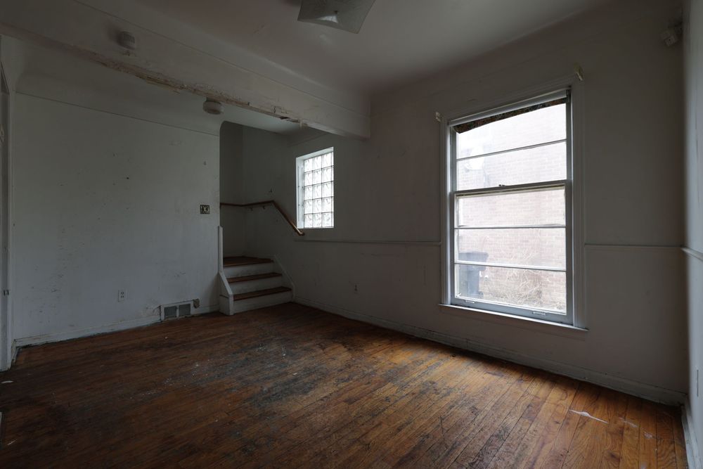 Empty room with hardwood floors, white walls, staircase, and two windows letting in natural light.