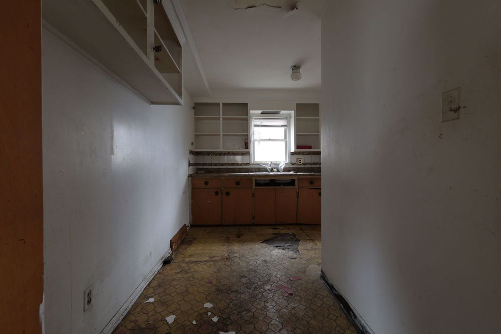 Abandoned kitchen with deteriorating floor, empty cabinets, and a window above the sink in a neglected home.