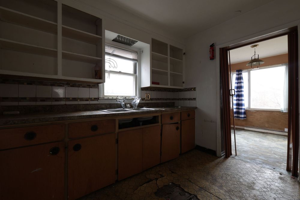 Abandoned kitchen with worn wooden cabinets, open shelves, dirty floors, and a window above the sink in a neglected home.