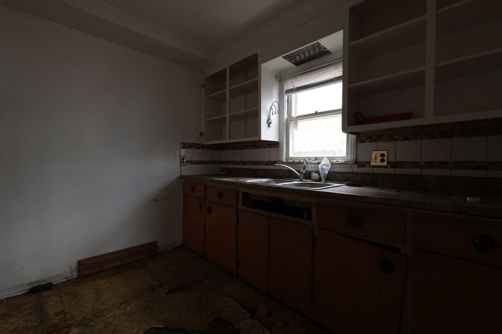 Abandoned kitchen with worn orange cabinets, empty shelves, a sink, and damaged flooring in a neglected interior.