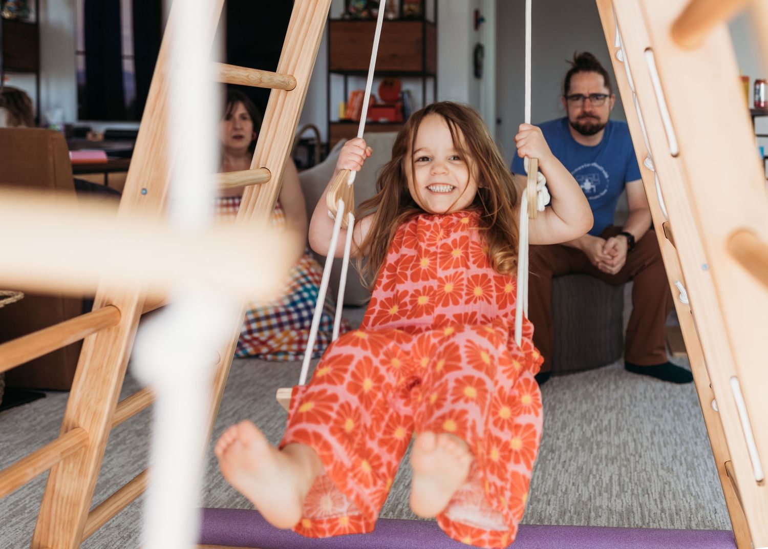 Young girl in red floral jumpsuit smiling on wooden indoor swing set with adults relaxing in background.