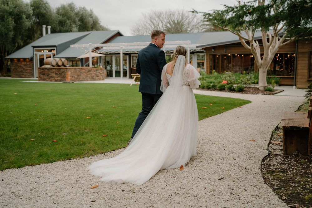 Elegant wedding ceremony with flowing white dress and veil trailing along a gravel pathway through lush garden grounds.
