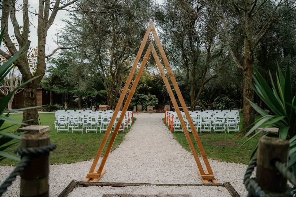 Wooden ceremony arch decorated with greenery stands in front of rows of white folding chairs in an outdoor garden setting.