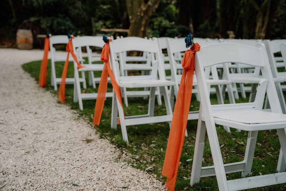 White folding chairs and orange ribbons line a garden path for an outdoor wedding ceremony setup.