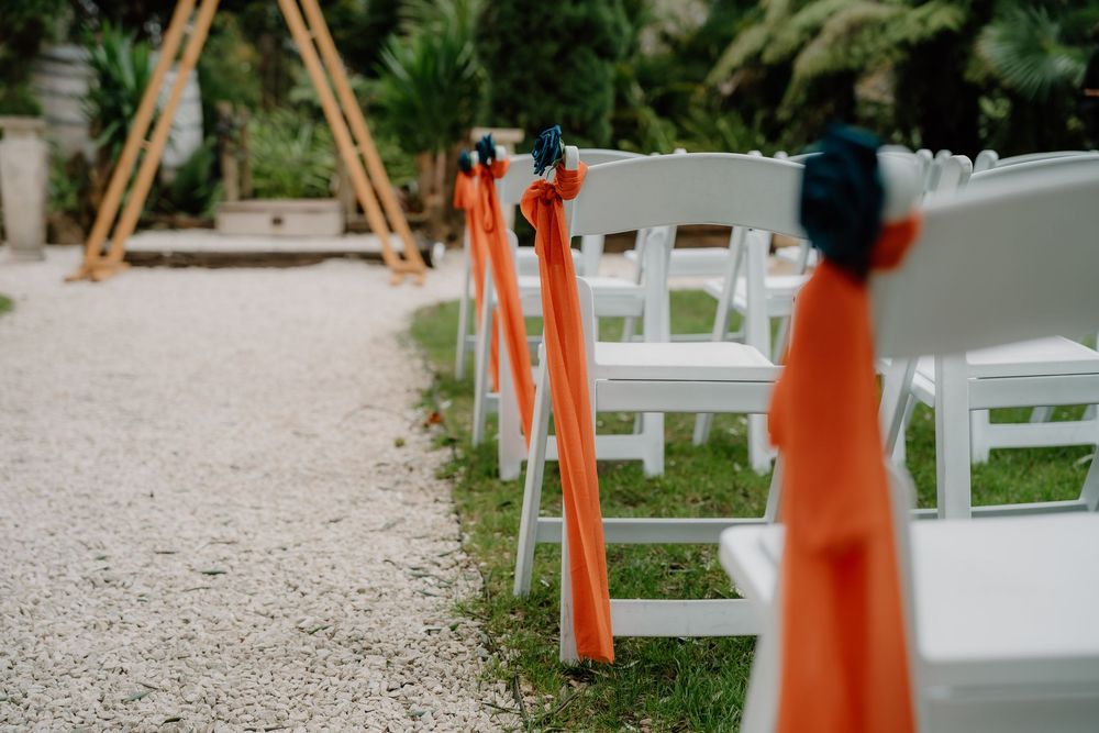 White folding chairs line a pebbled outdoor pathway with orange traffic cones marking the aisle at a wedding ceremony.