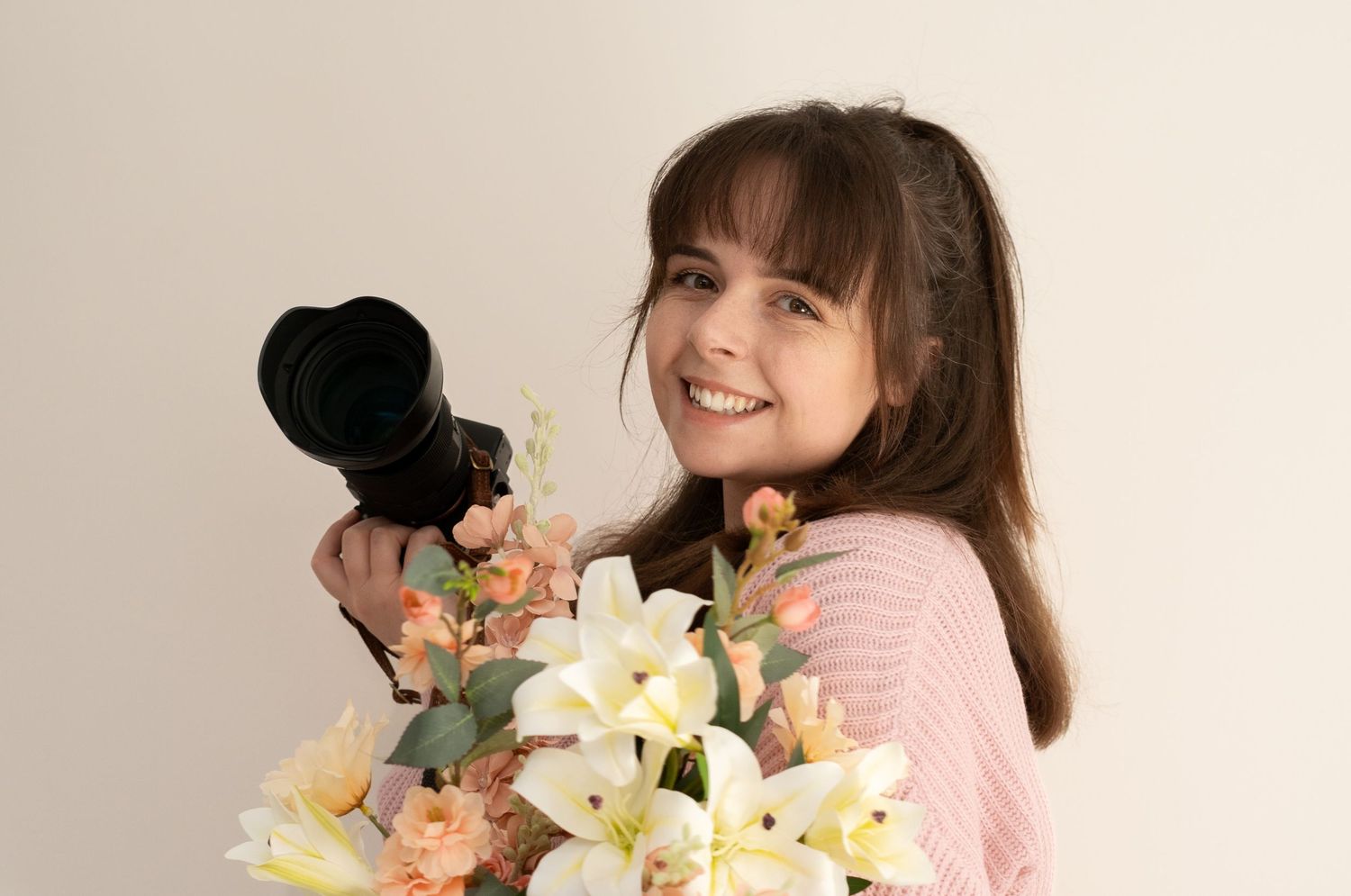 Smiling young woman holding a camera and large bouquet of white and peach flowers in a pink sweater.