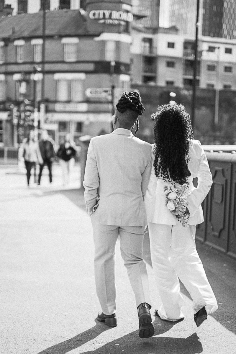 Black and white photo of a couple in white wedding attire walking hand in hand on a city bridge.