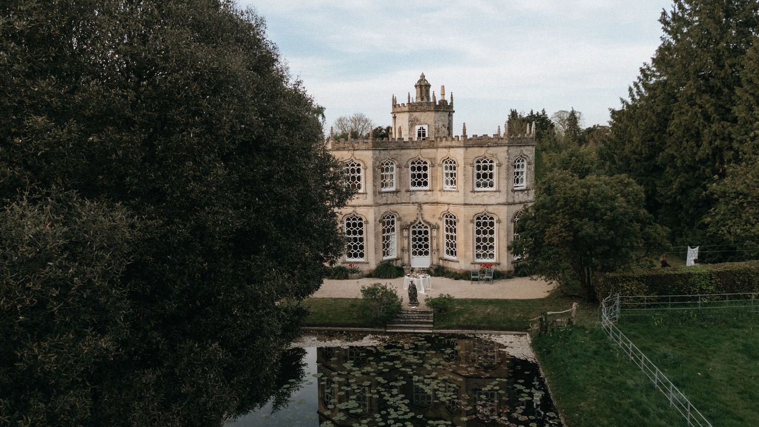 Aerial view of a grand Georgian manor house surrounded by lush trees and a ornamental pond in manicured grounds.