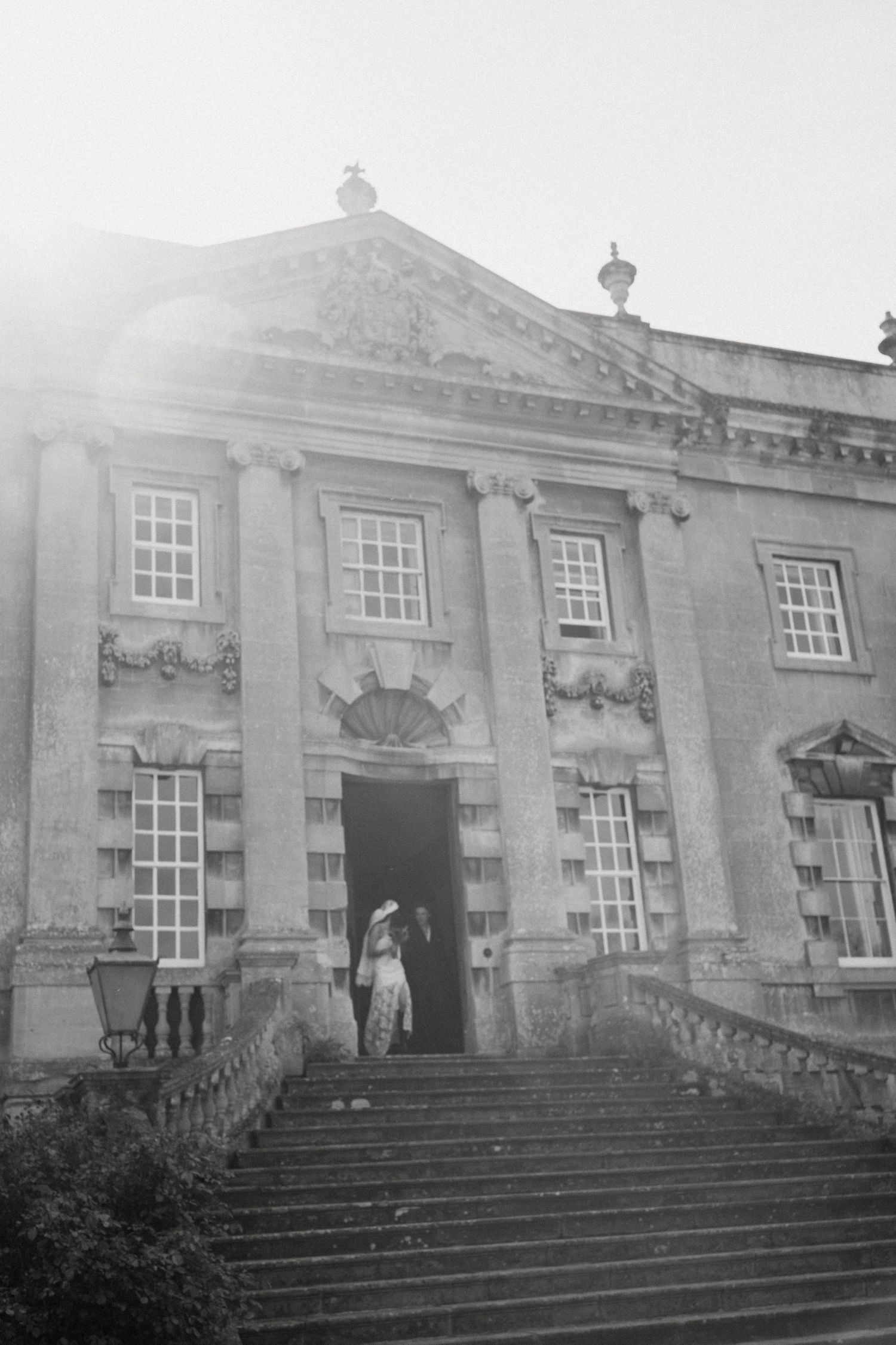 Black and white photo of a couple on grand stone steps of a classical Georgian mansion with ornate columns.