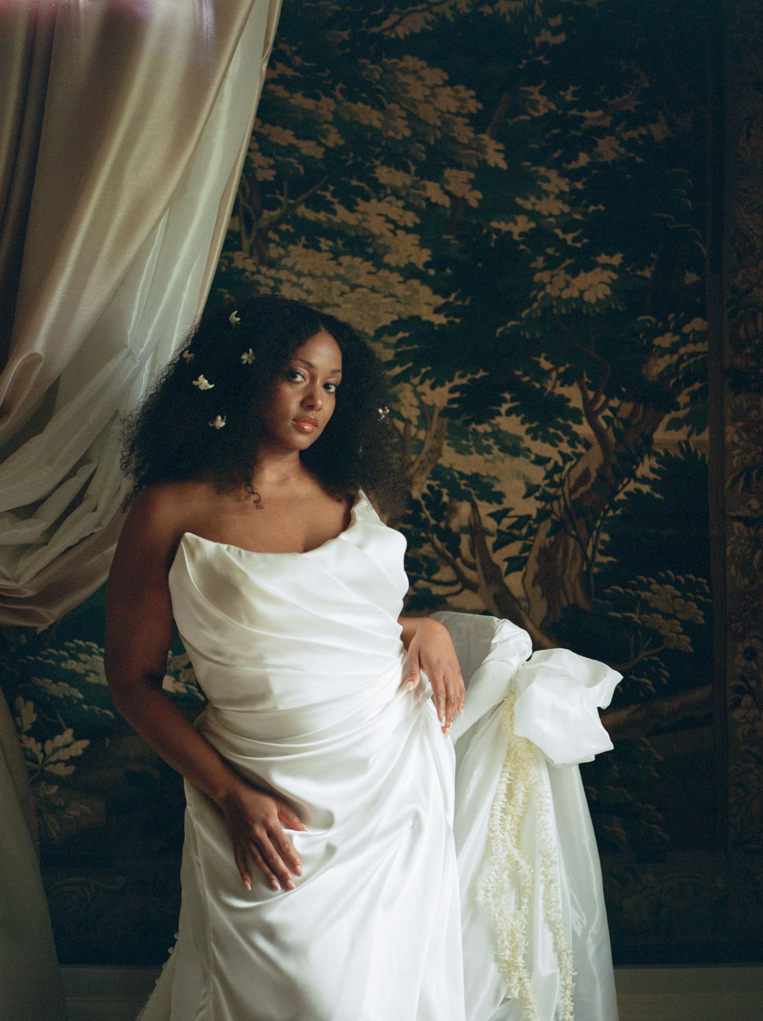 A woman in an elegant white draped gown stands before a tapestry backdrop with floral hair accessories.