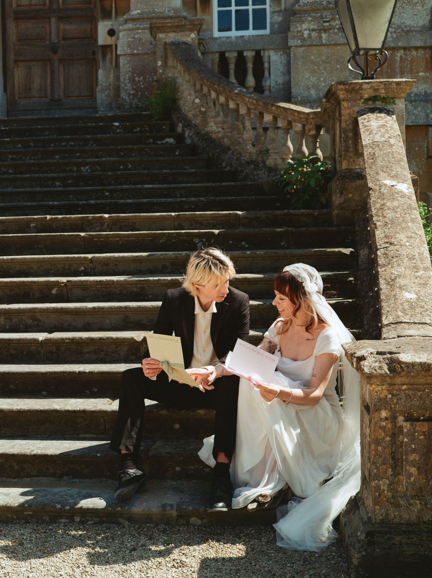 Bride and groom sitting on stone steps of a grand estate, reading wedding vows together in bright sunlight.