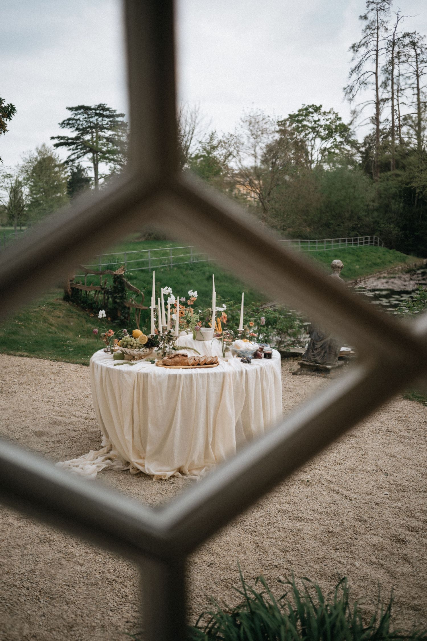 Elegant outdoor wedding table with white draping, candles, and floral arrangements viewed through a diamond-paned window.