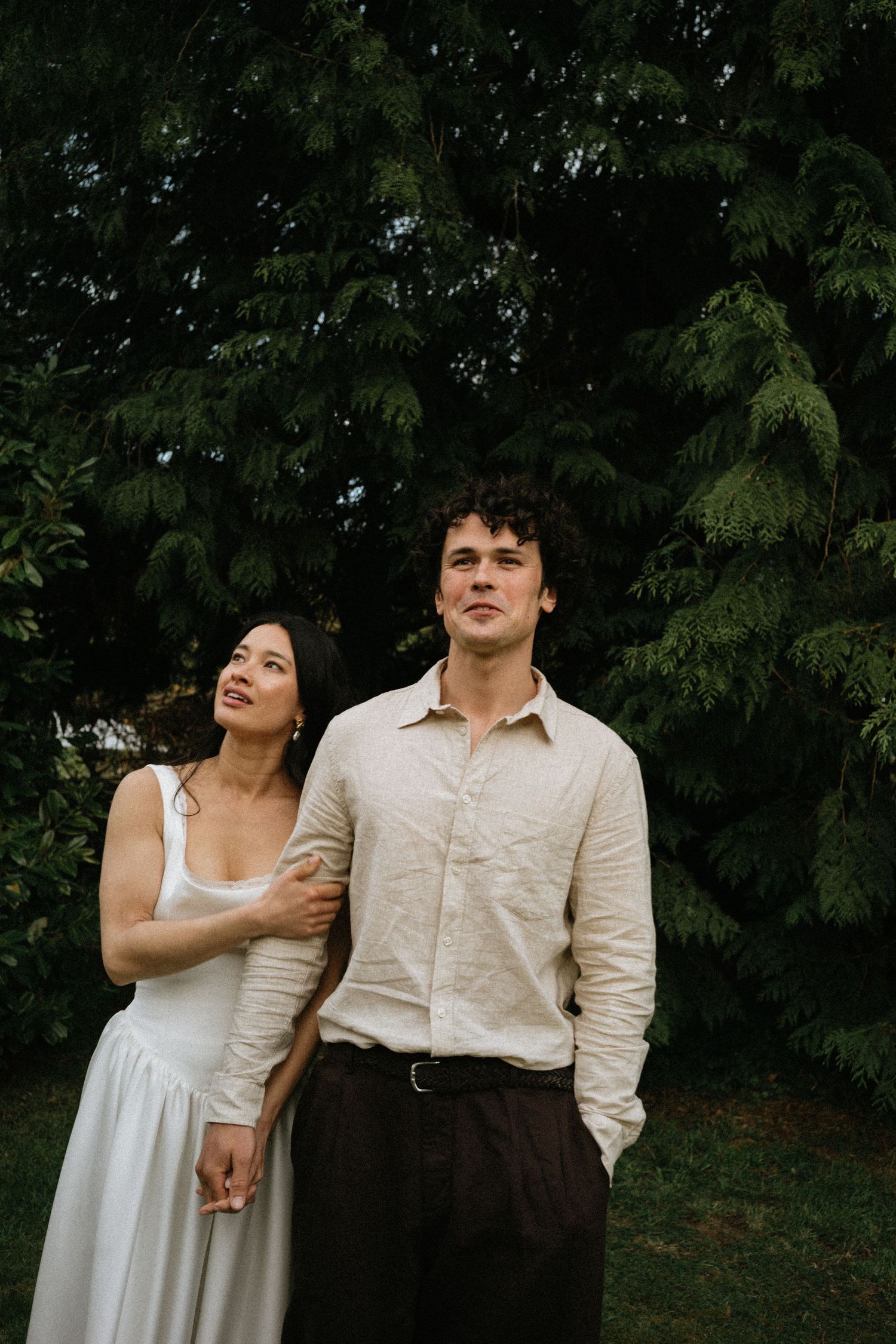 A couple in elegant attire stands outdoors before lush green trees, the woman in white dress holding the man's arm.