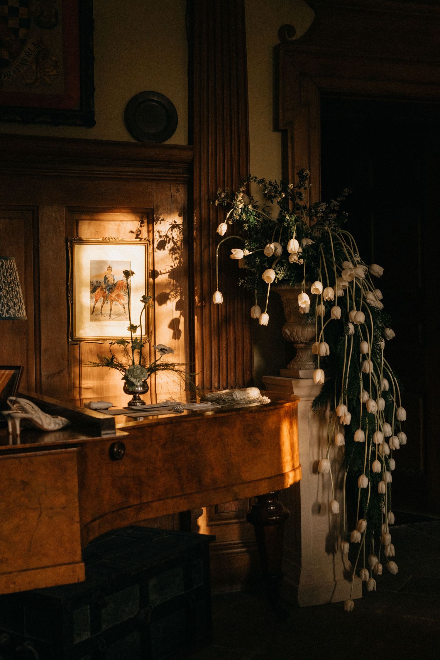 Elegant dark interior with cascading white tulips in urn vase beside antique wooden dresser with framed equestrian art.
