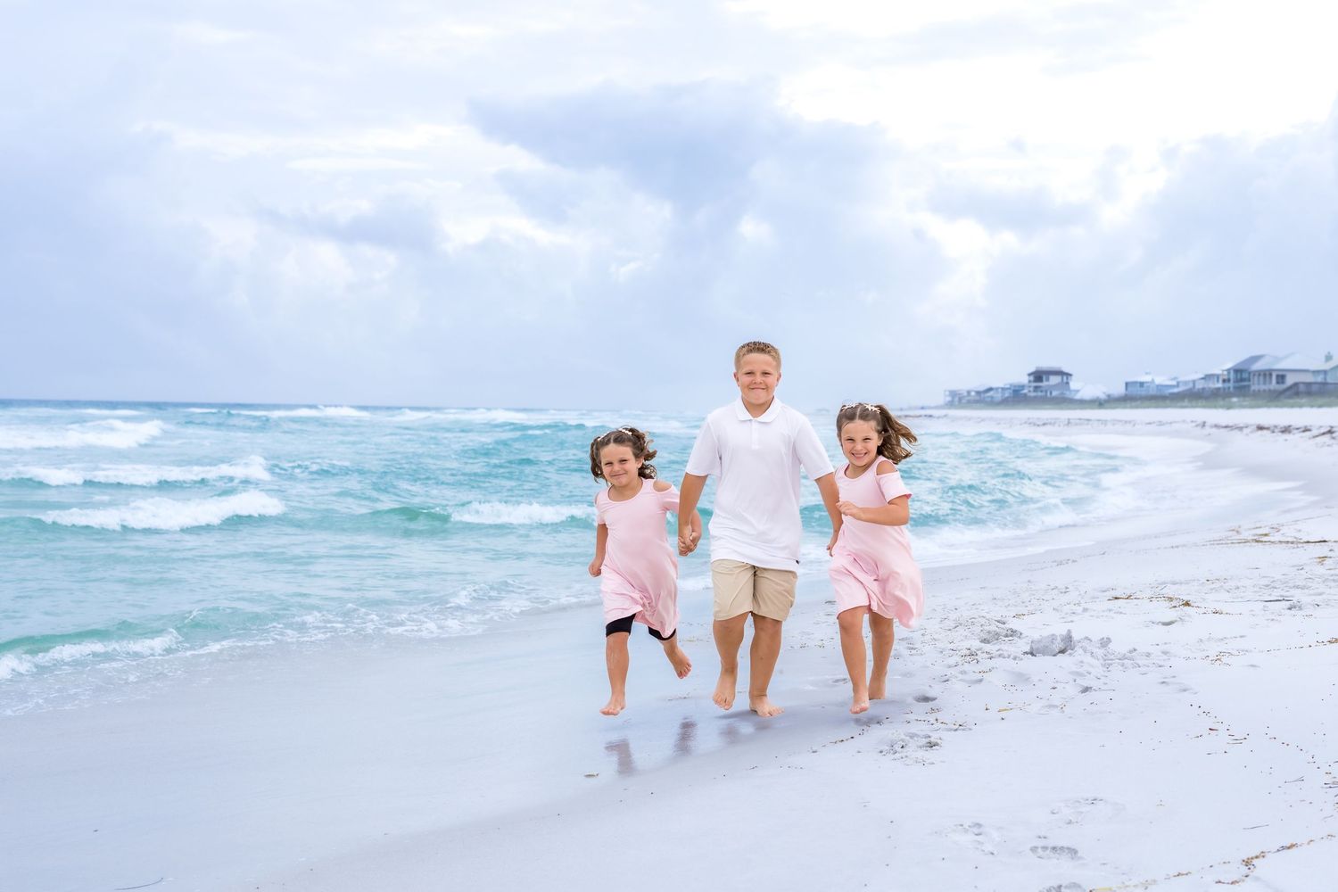 A mother hugs her two young daughters in pink dresses on a sandy beach with turquoise waves behind them.