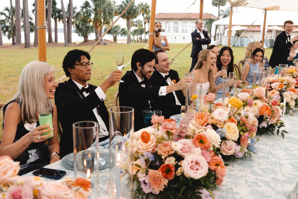 Wedding reception guests laughing at head table adorned with colorful pink and orange floral arrangements under outdoor tent.