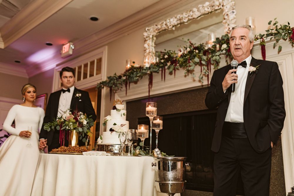 Father of the groom gives a heartfelt wedding toast at elegant reception with floral fireplace backdrop.