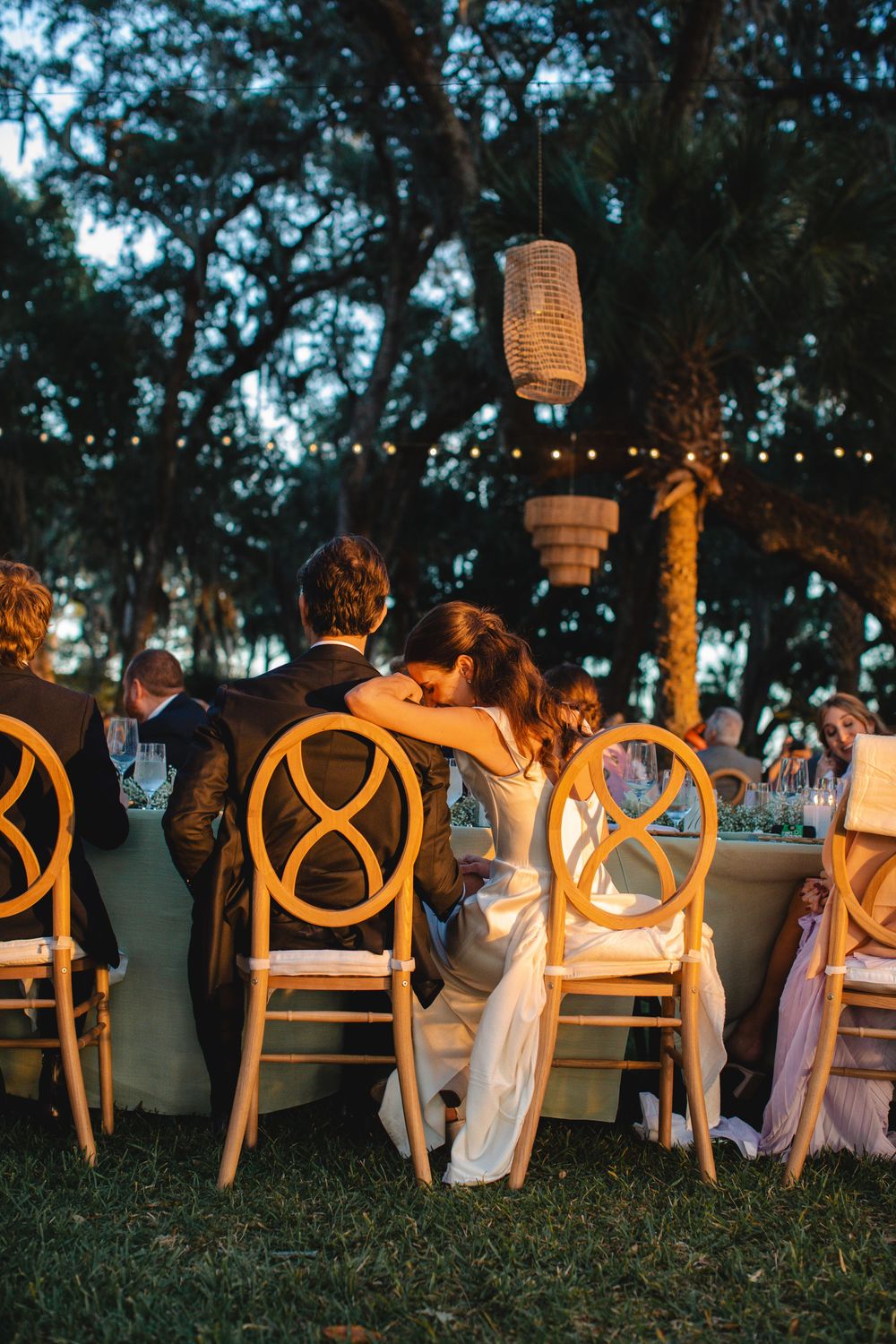 Bride and groom share a romantic kiss at an outdoor evening wedding reception under string lights and hanging lanterns.