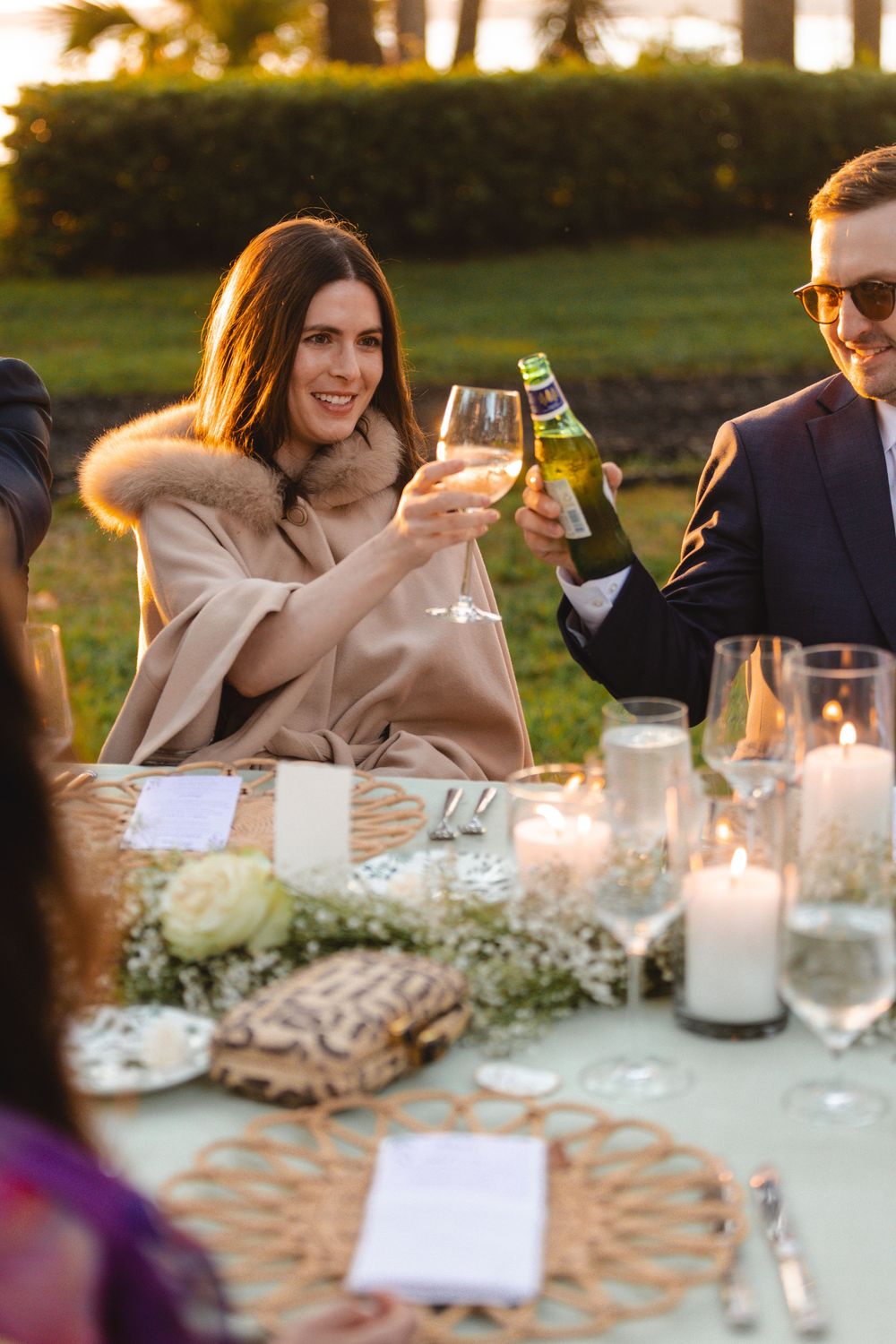 Elegant outdoor dinner party toast with woman in beige fur-trimmed cape raising wine glass at candlelit table.