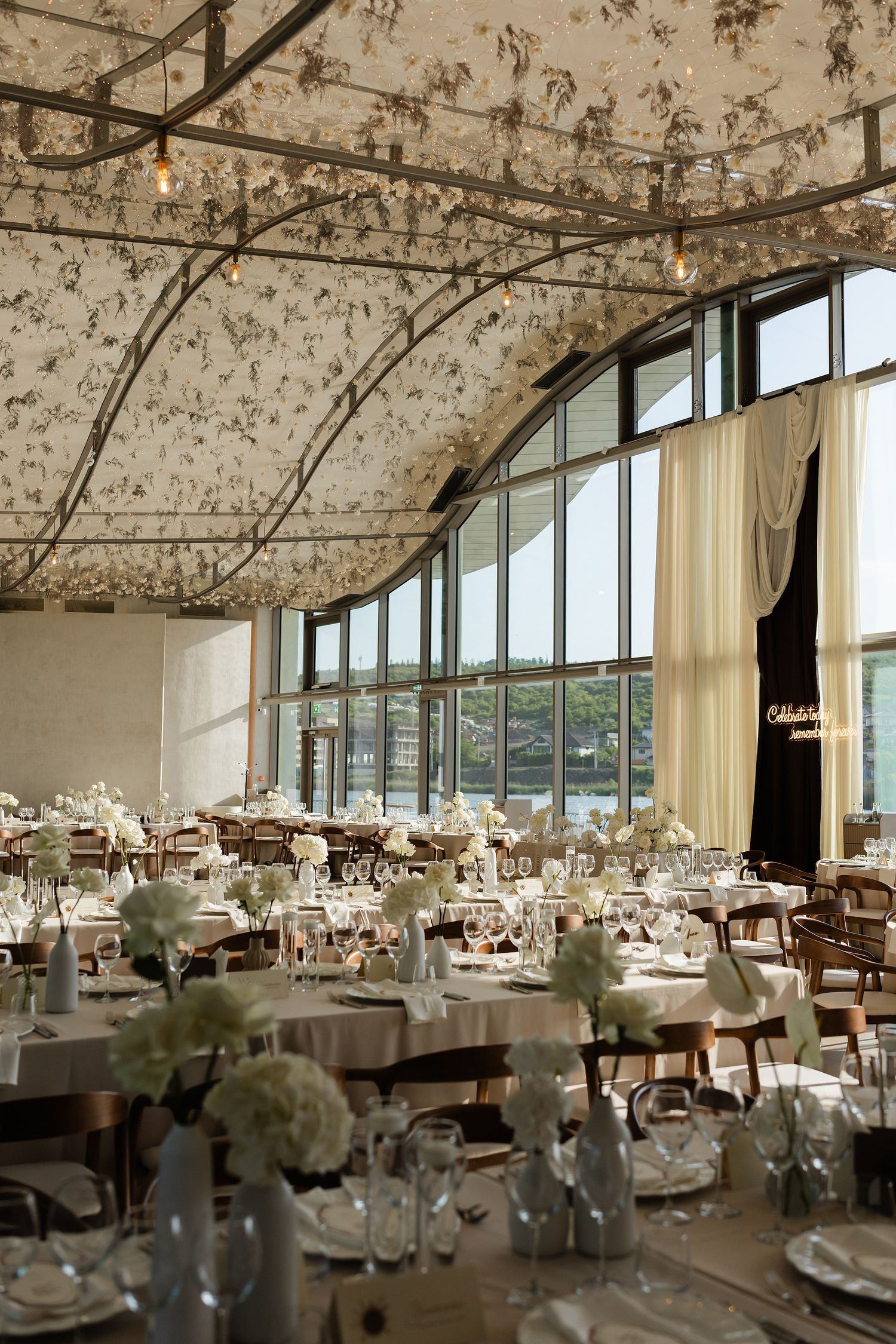 Elegant wedding reception hall with floral ceiling decor, white tablecloths, hydrangea centerpieces, and large arched windows.