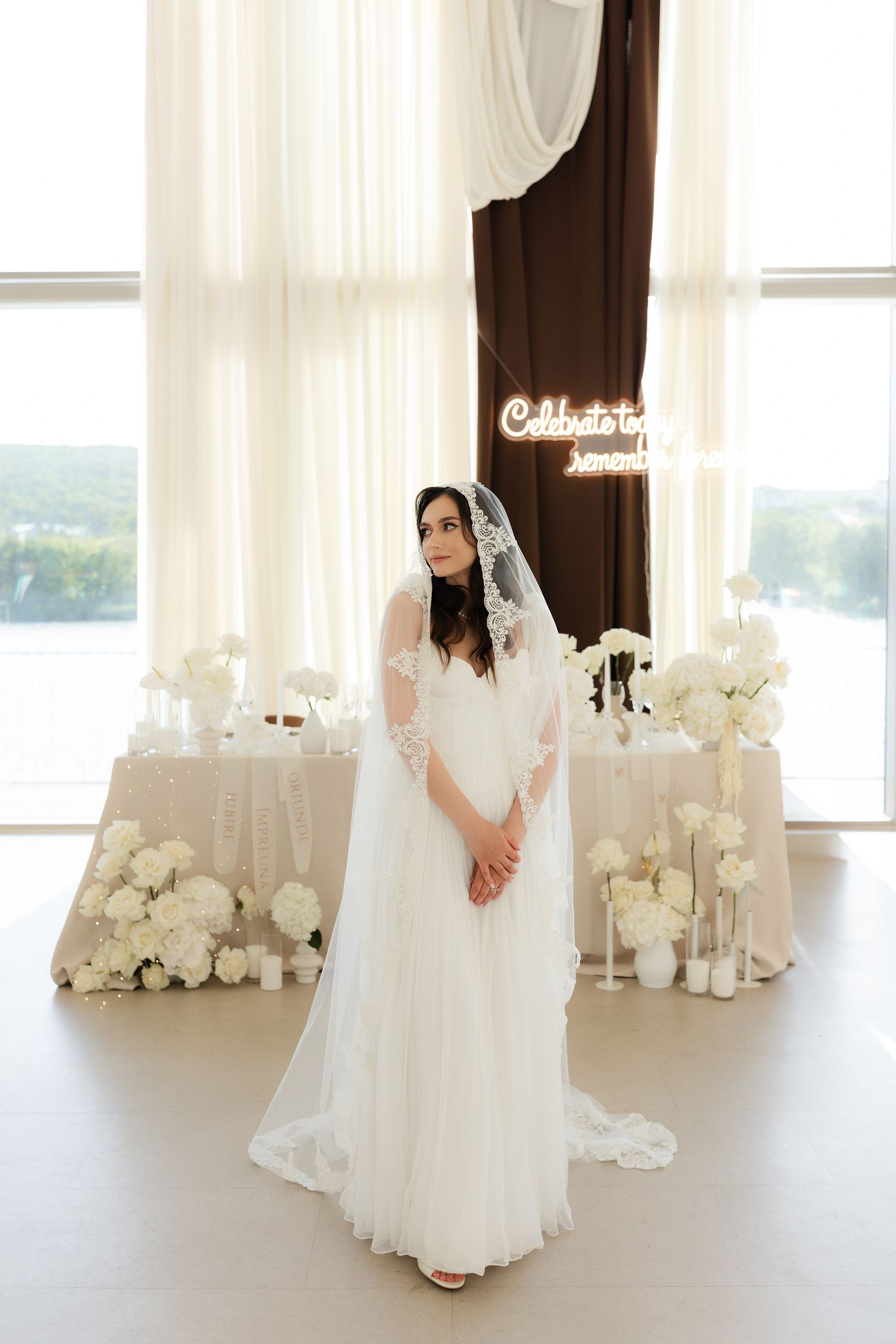 Elegant bride in white lace veil and gown stands before floral arrangements and neon sign at wedding venue.