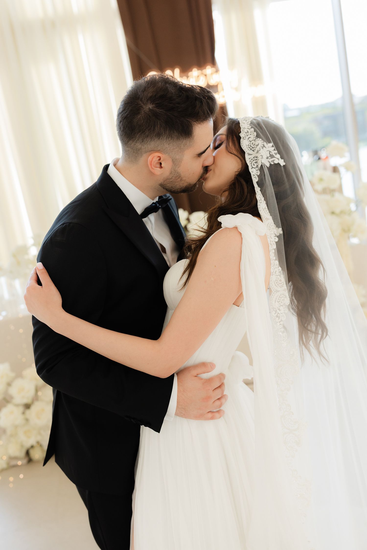 A bride and groom share a romantic first kiss at their wedding reception, dressed in elegant white gown and black tuxedo.