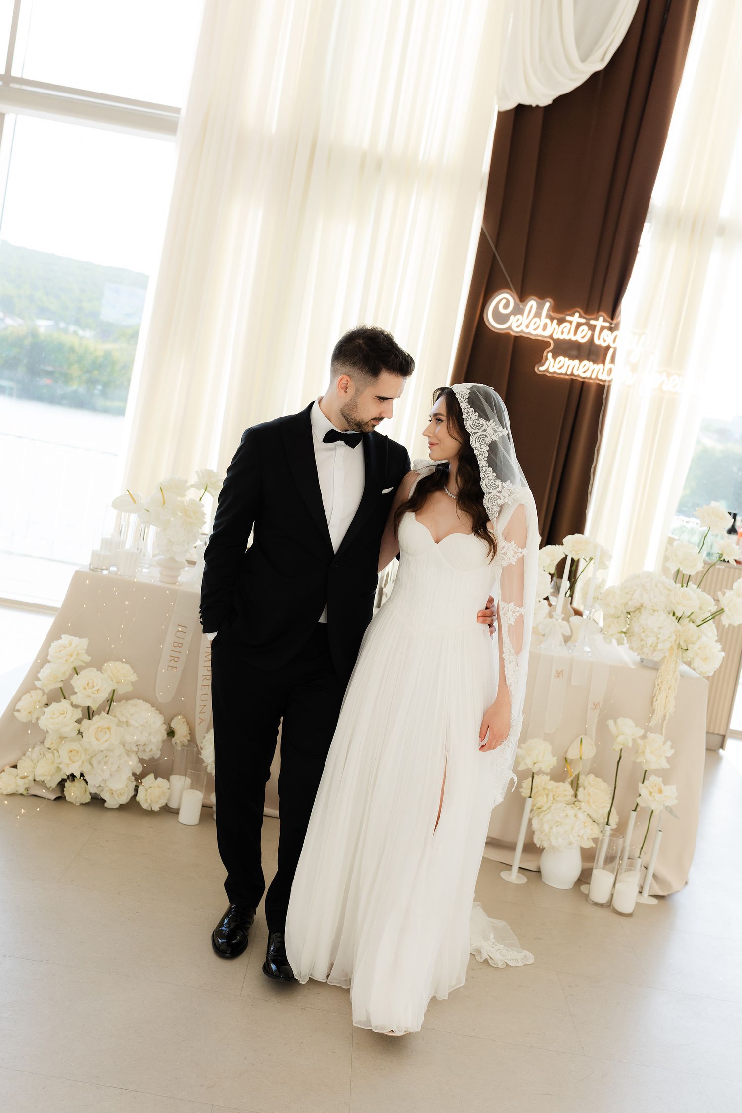 Elegant bride and groom posing together at their wedding reception with white floral arrangements and neon sign.