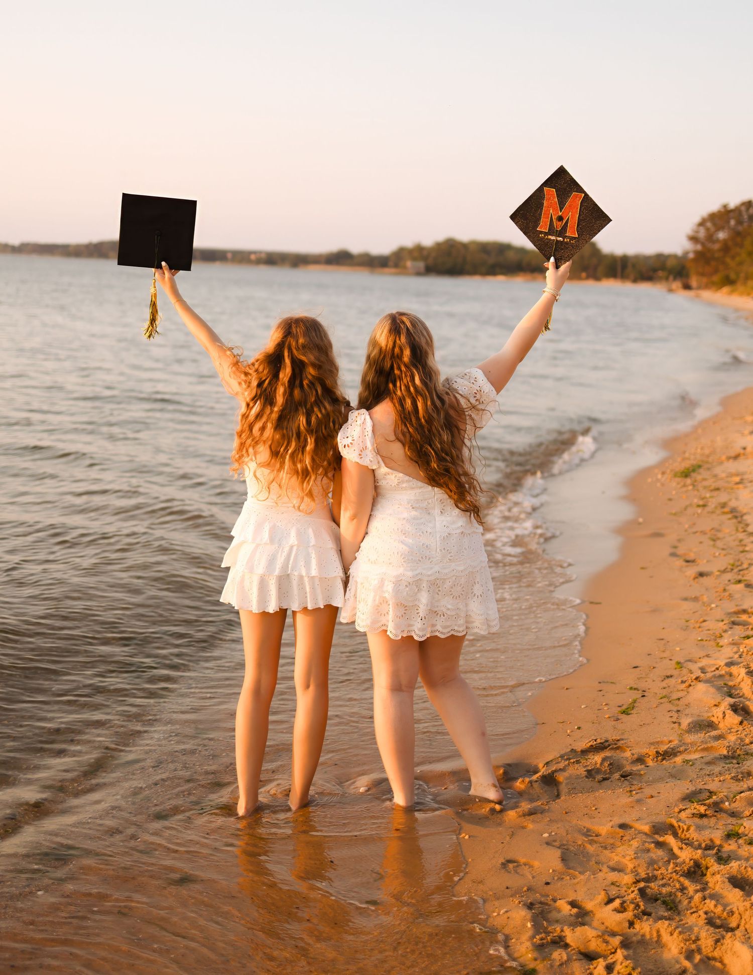 Two graduates with long curly hair hold black mortarboards with letter M near a beach at sunset.