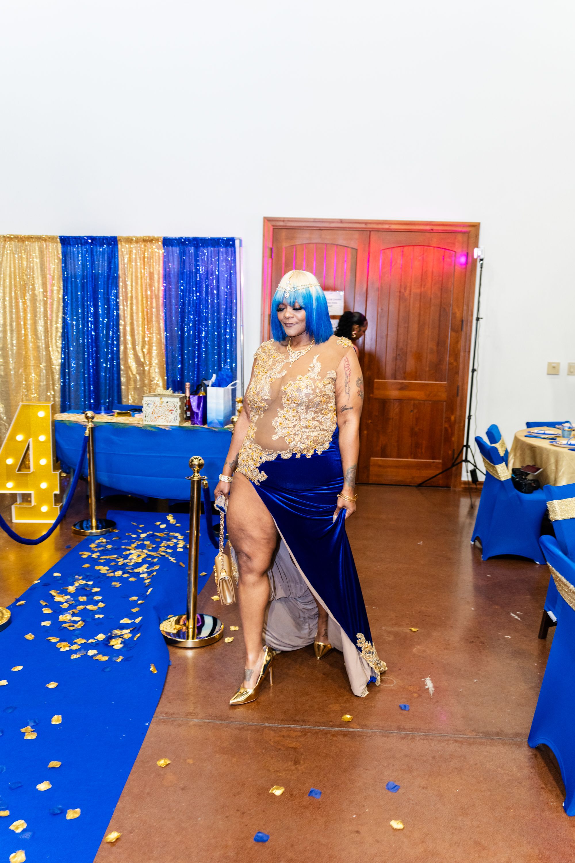 Woman in blue wig and gold lace gown walks blue carpet at elegant event with gold decorations.