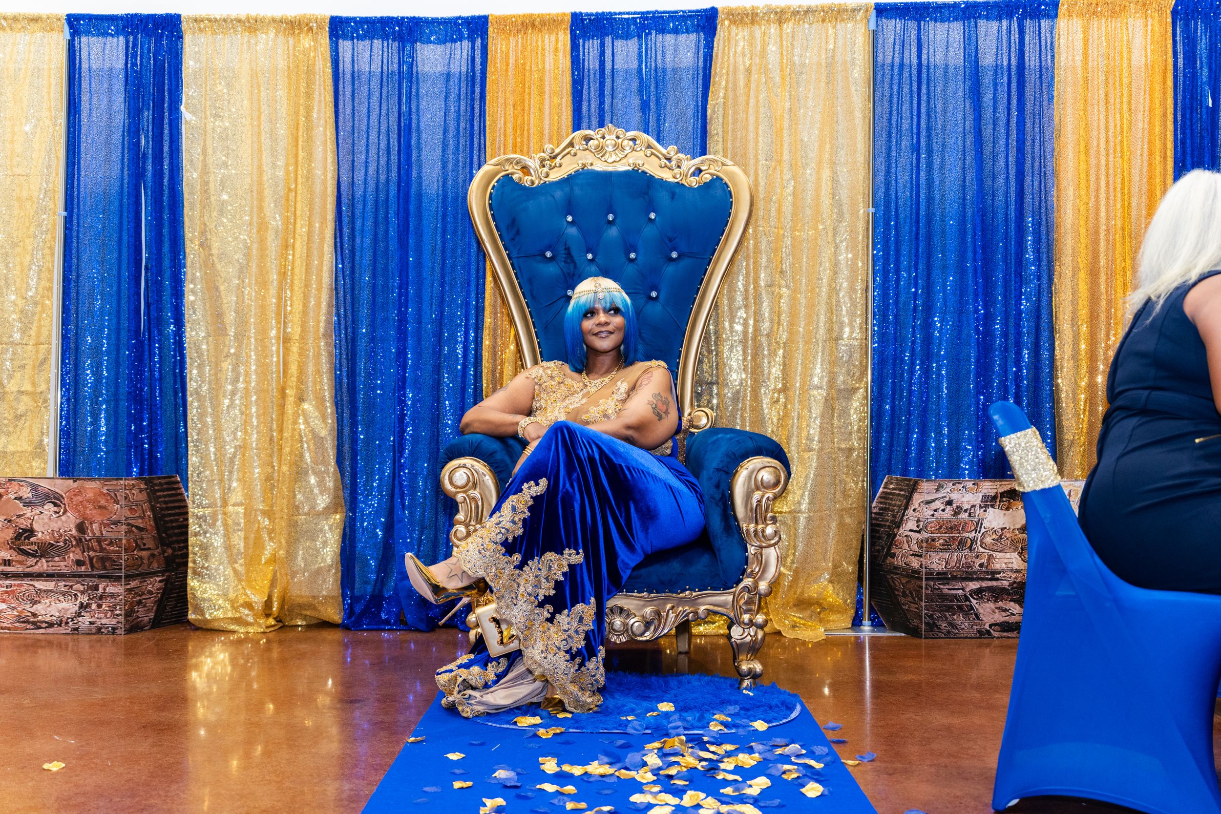 Woman in blue gown seated on ornate gold throne chair with royal blue and gold sequin backdrop.