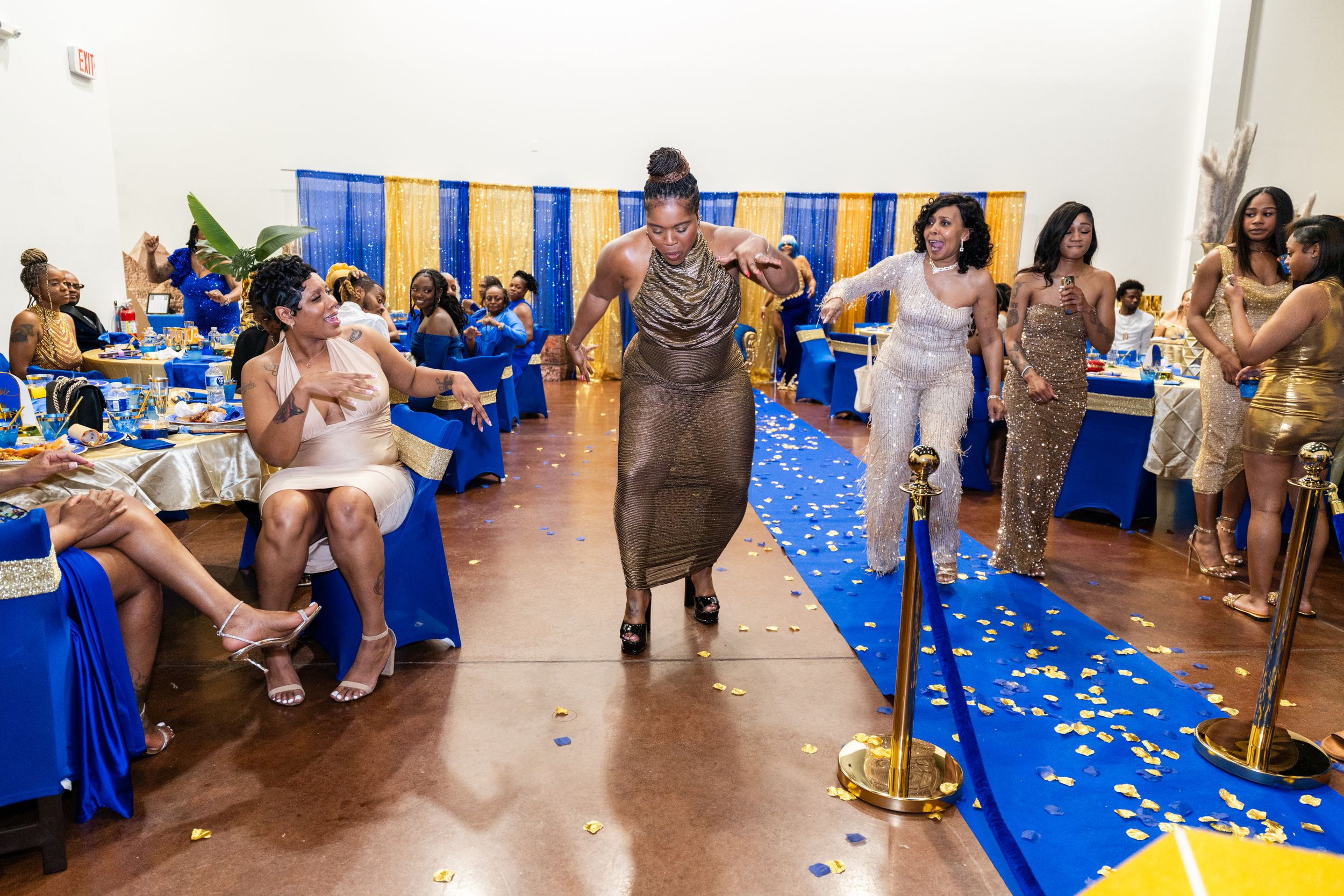 Woman in gold dress walks runway at elegant blue and gold banquet event with guests seated at decorated tables.