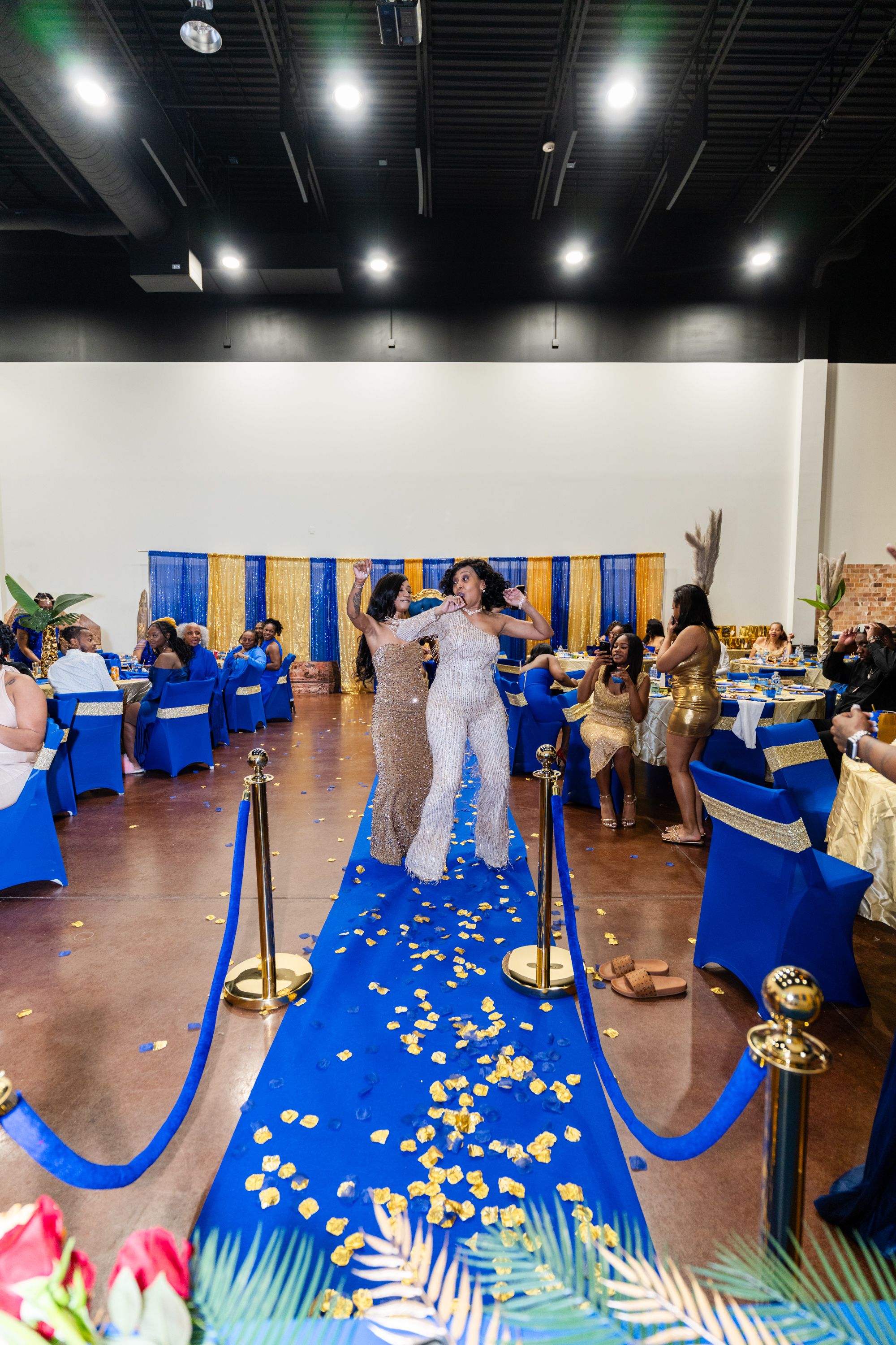 Woman walks blue carpet runway at elegant royal blue and gold themed banquet celebration event.