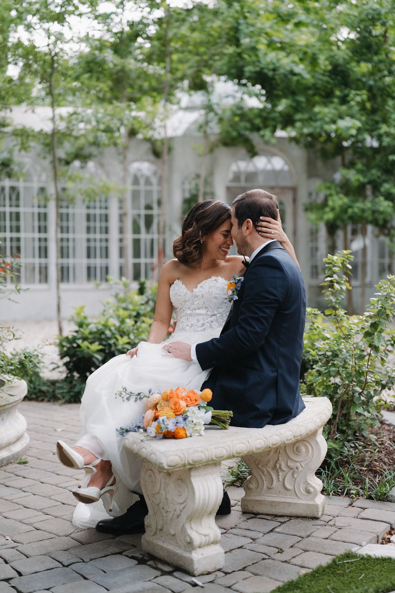 Bride and groom sitting on a stone bench in a lush garden, sharing a romantic moment with an orange and blue bouquet.