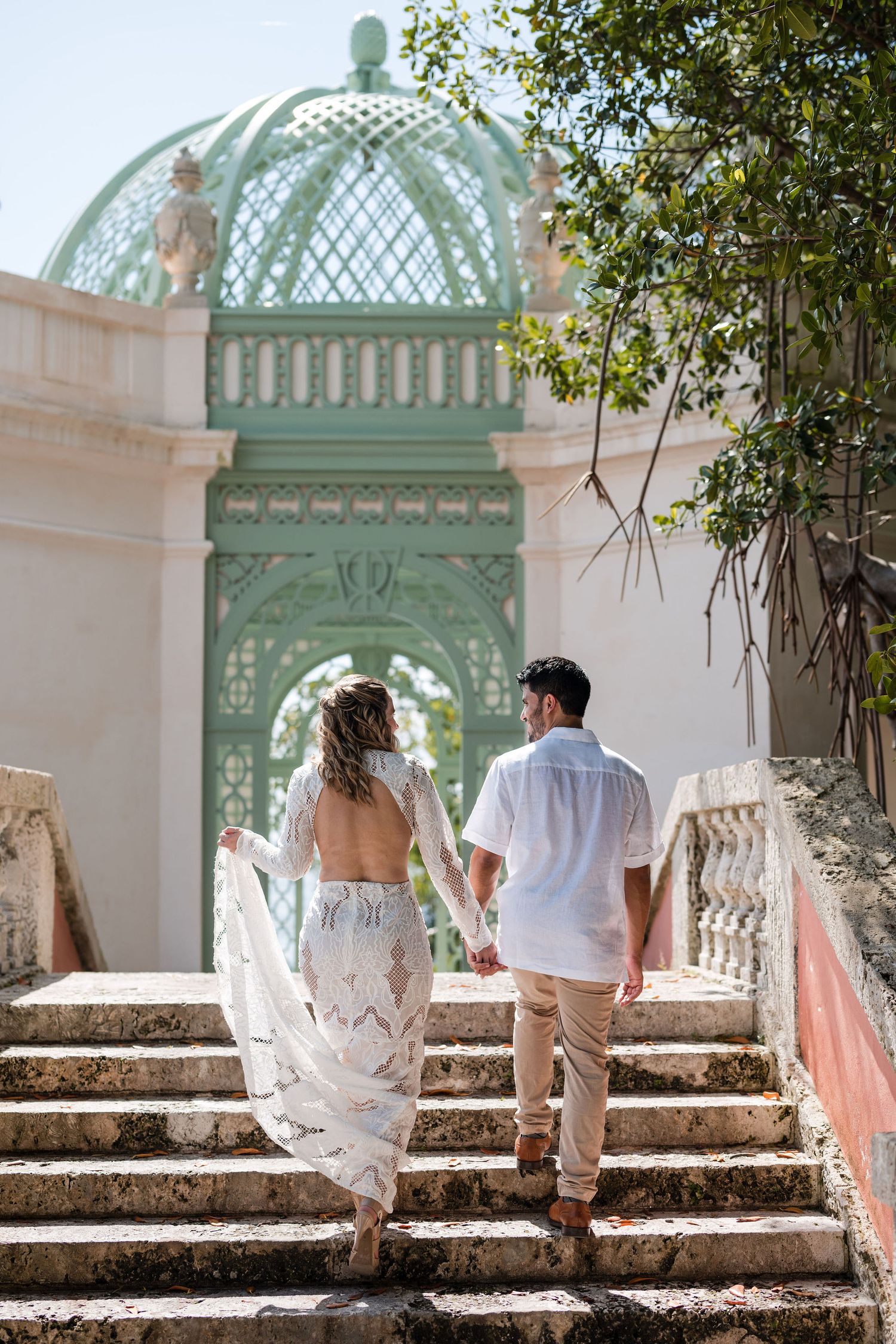 Bride and groom walk hand in hand up stone steps at Vizcaya Gardens, framed by elegant architecture and a historic green pavilion.