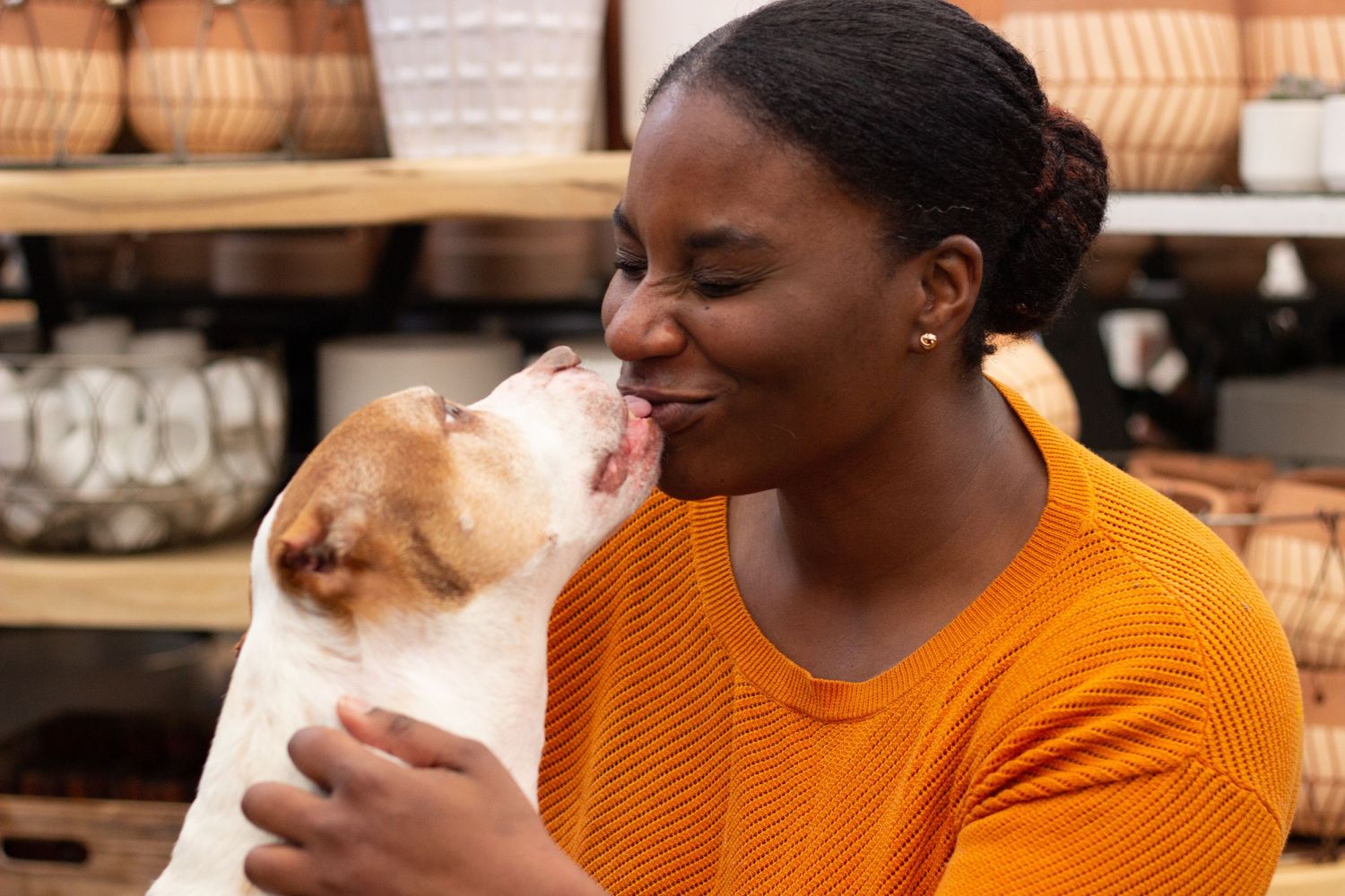 Person in orange sweater receiving affectionate kiss from small dog in retail setting.