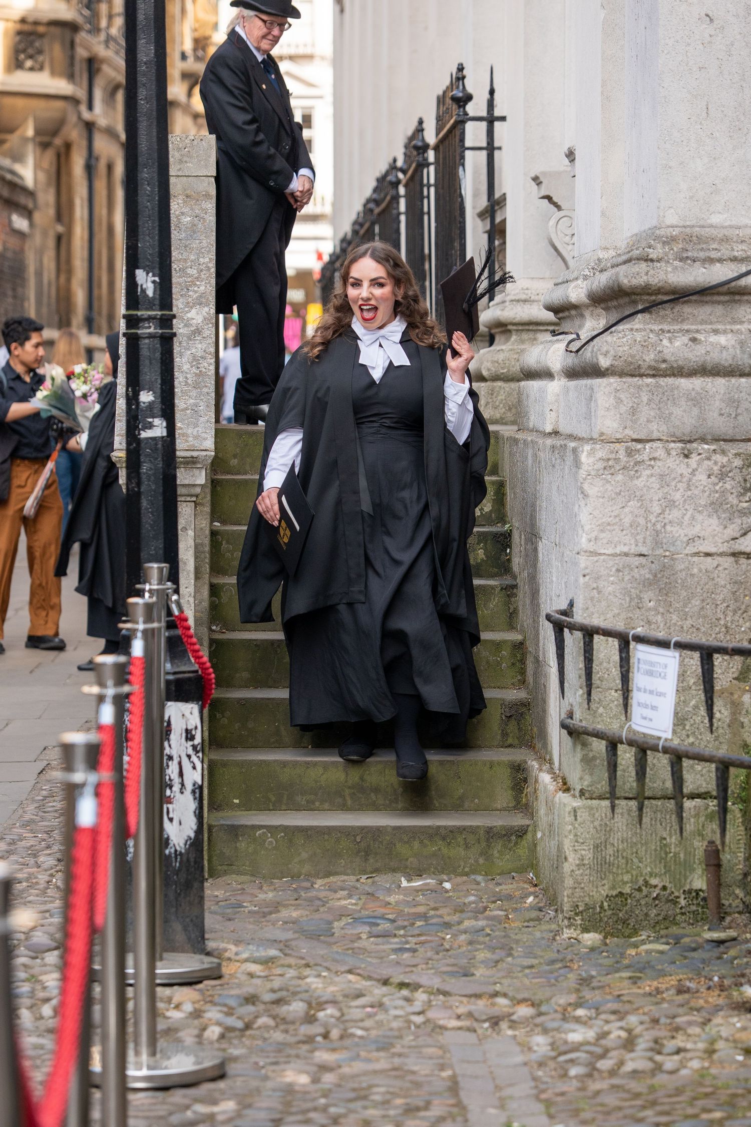 Excited graduate in black academic gown holding diploma and mortarboard on cobblestone path.