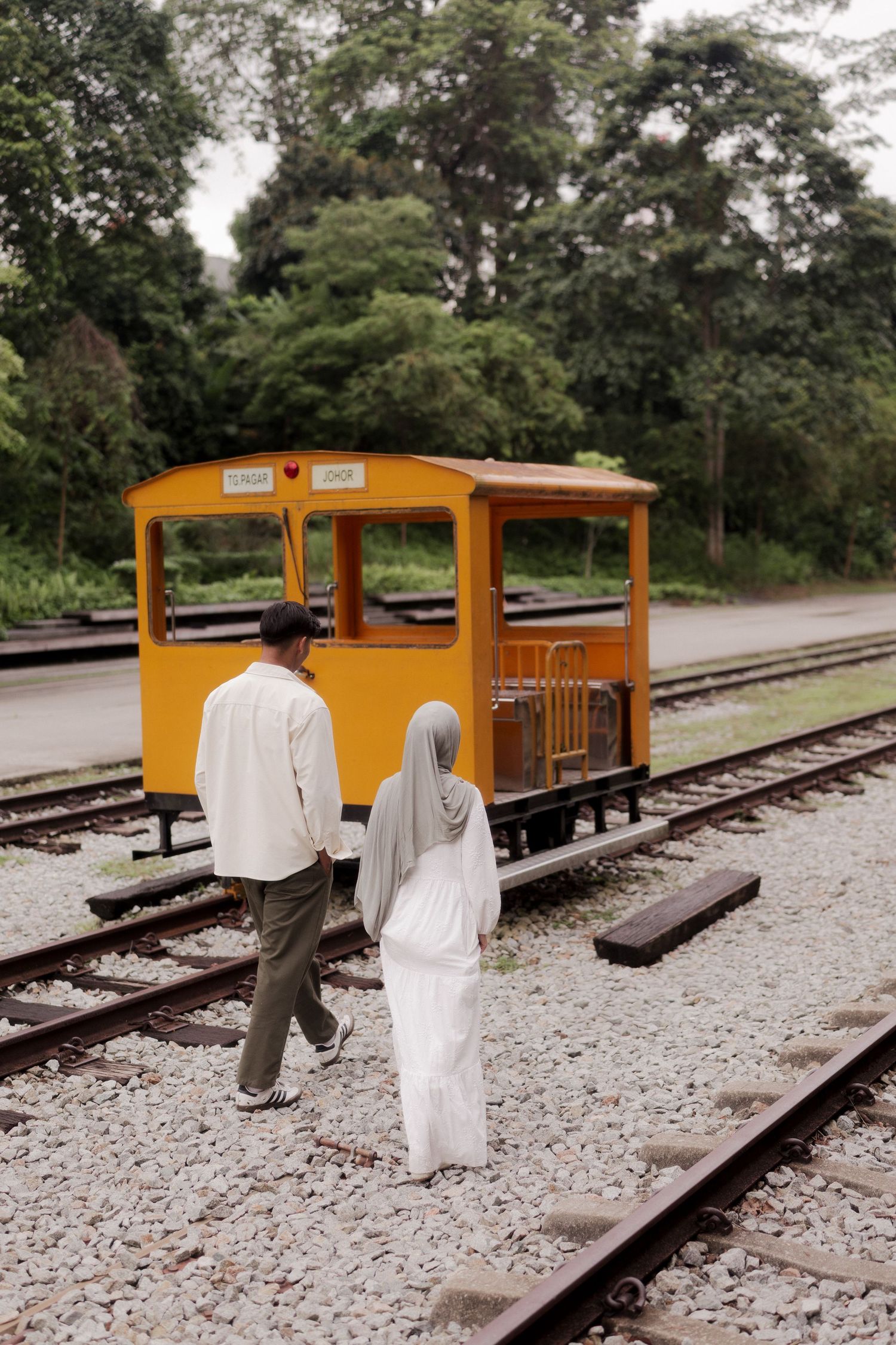 Couple walking hand in hand toward a yellow vintage train car on railroad tracks surrounded by lush green trees.