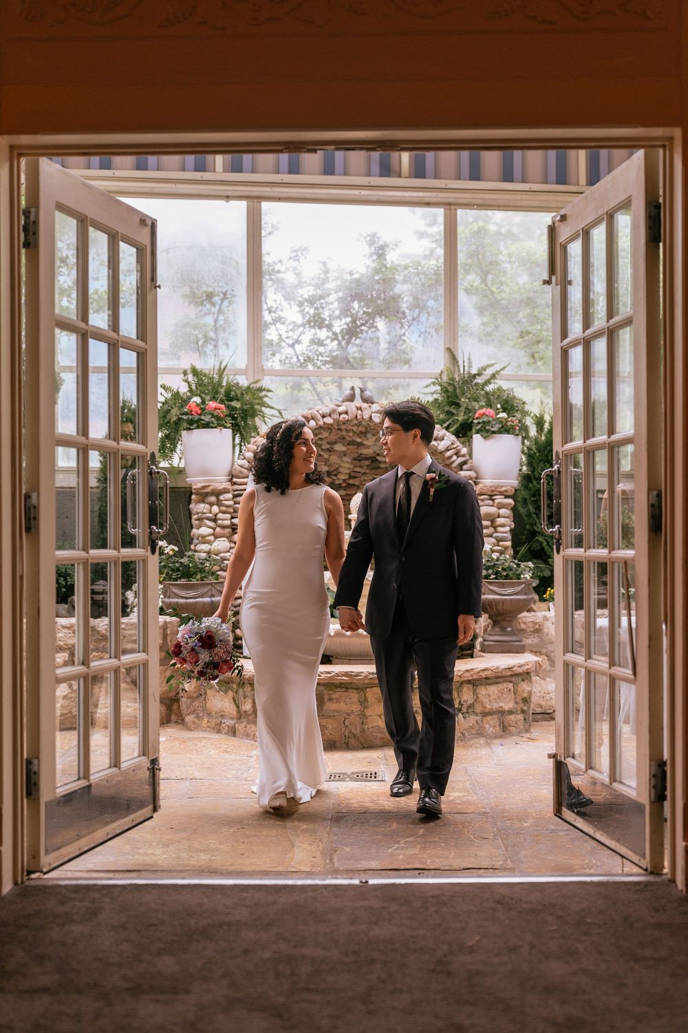 Bride and groom walk hand-in-hand through open French doors at an elegant garden wedding venue.
