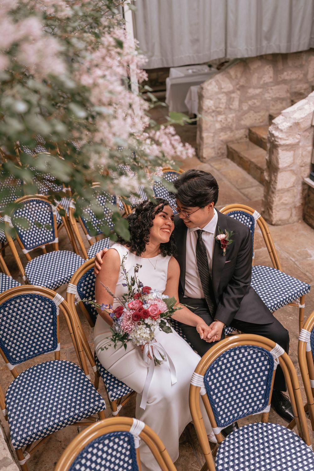 Bride and groom sitting among blue bistro chairs, surrounded by pink blossoms, sharing a tender moment with a bridal bouquet.