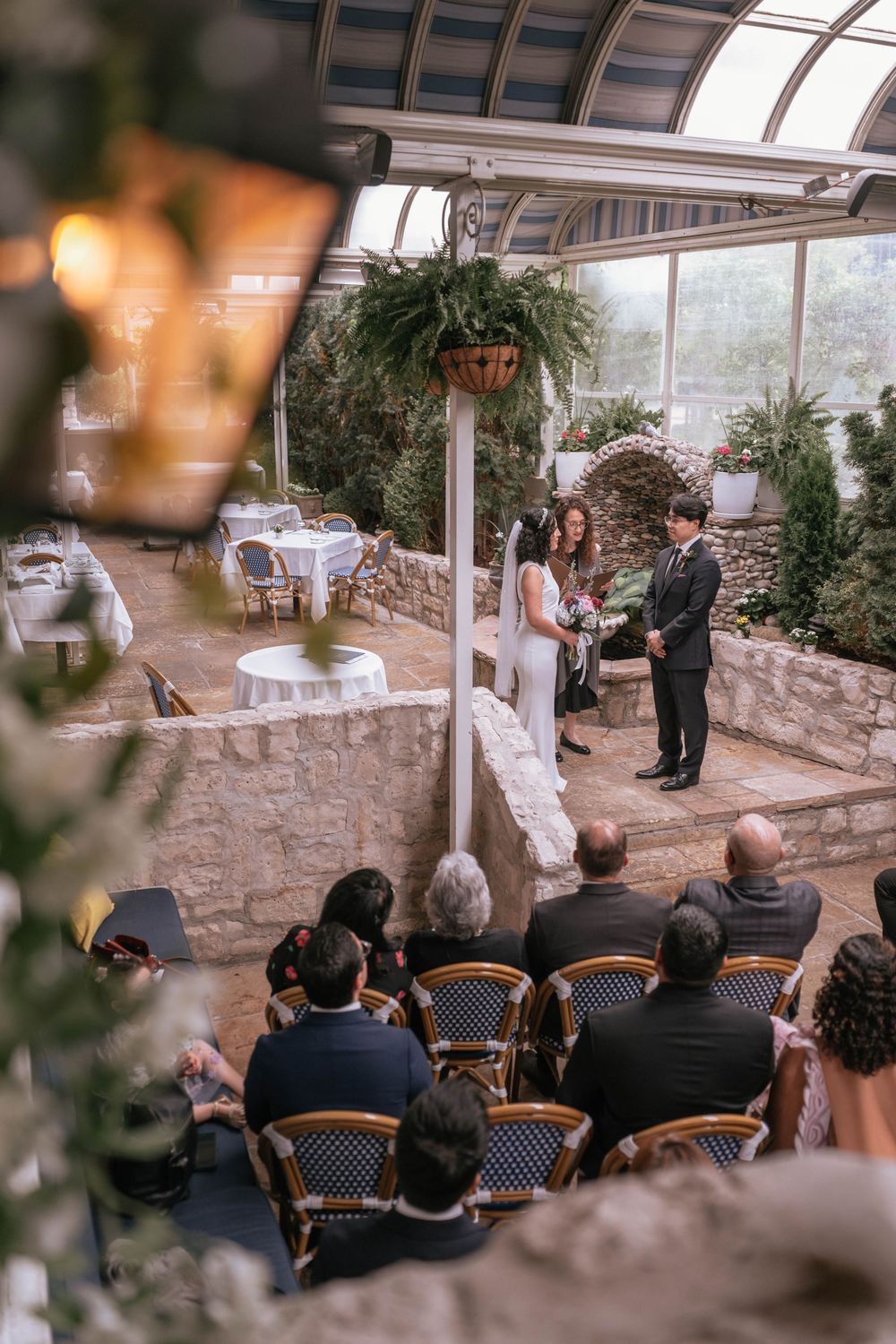 A wedding ceremony in a greenhouse venue with guests seated as a couple exchanges vows before an officiant amid lush greenery.