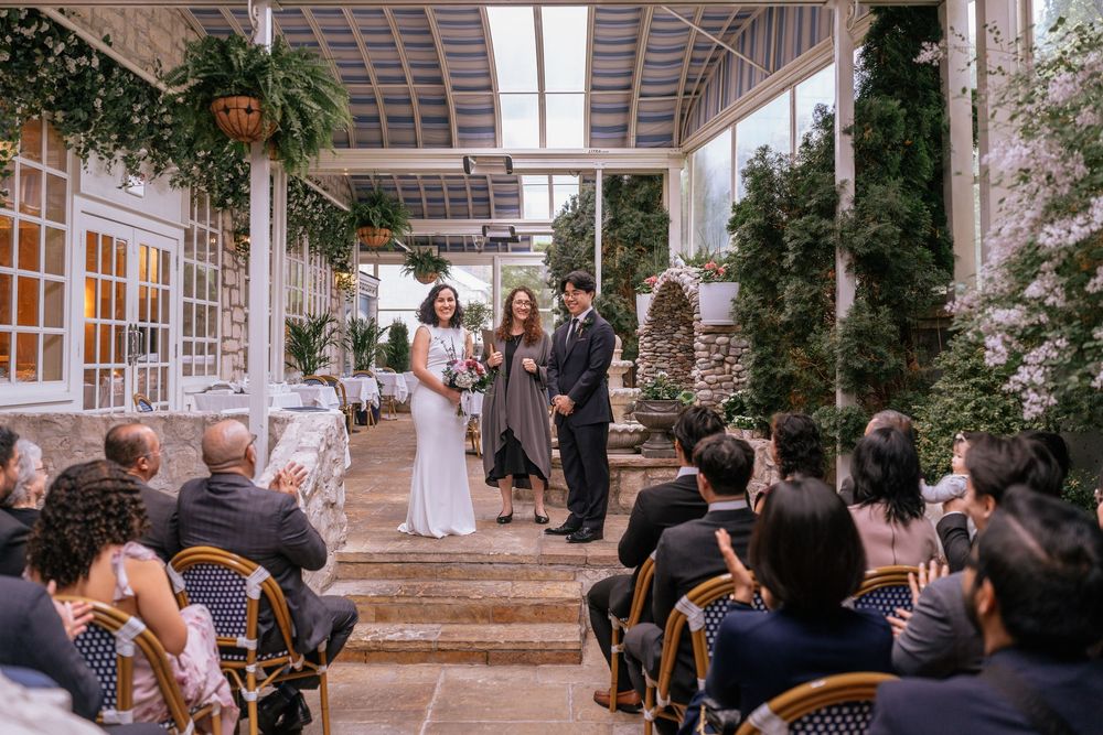 Elegant indoor wedding ceremony with guests seated in black chairs, bride and groom at altar surrounded by lush greenery.