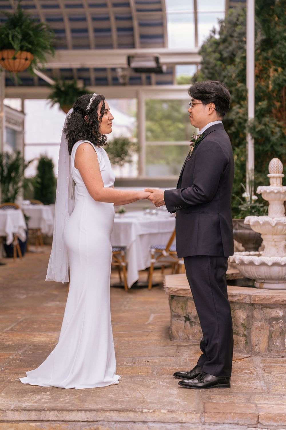 Bride and groom holding hands during outdoor wedding ceremony near stone fountain and greenhouse venue.