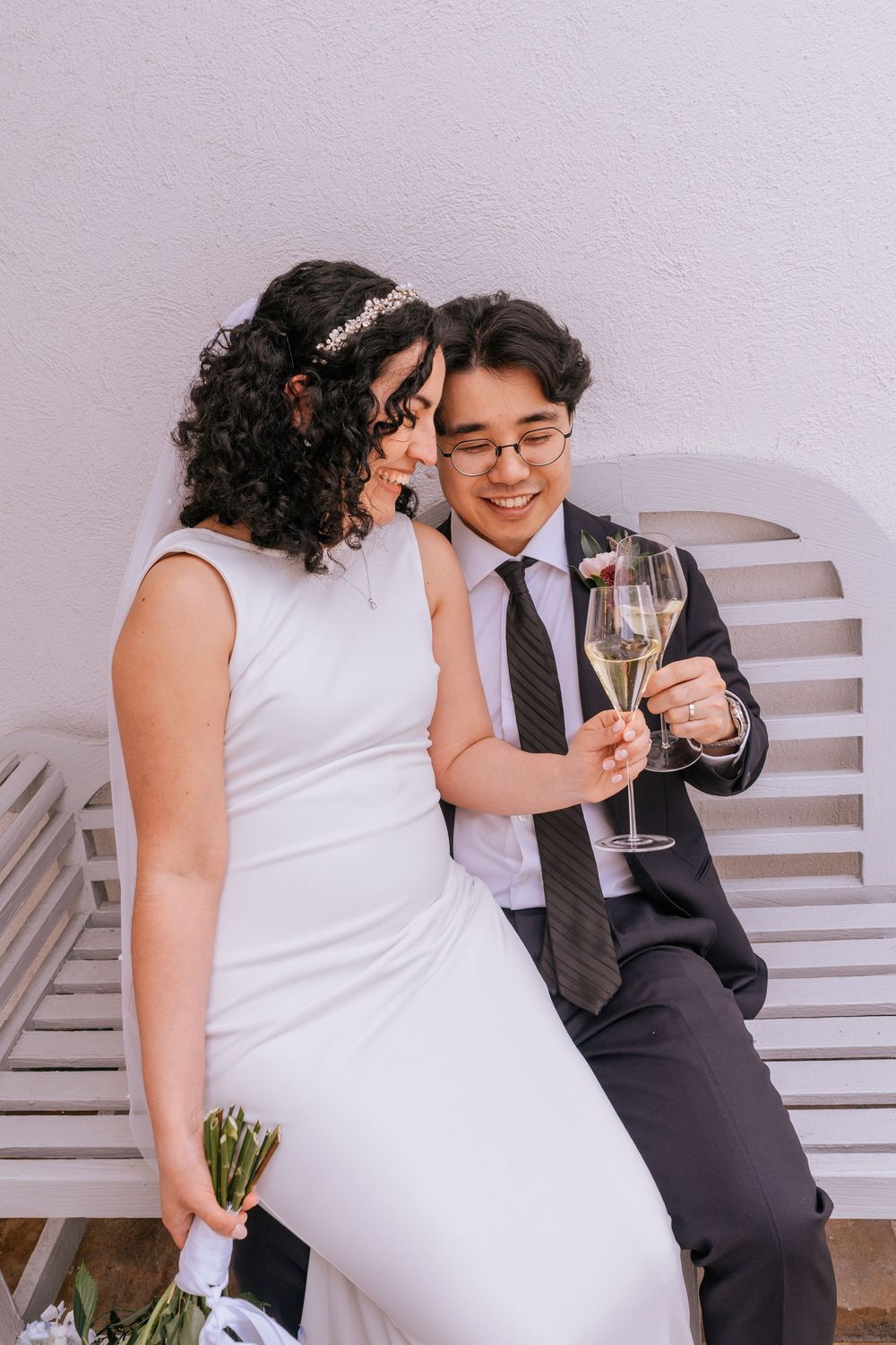 Newlyweds toast with champagne glasses while sitting on a white bench, smiling joyfully.
