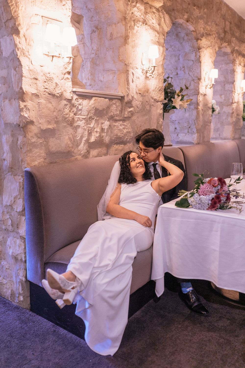 Bride in white dress reclines on groom's lap in cozy stone-walled venue, sharing tender moment at wedding reception.