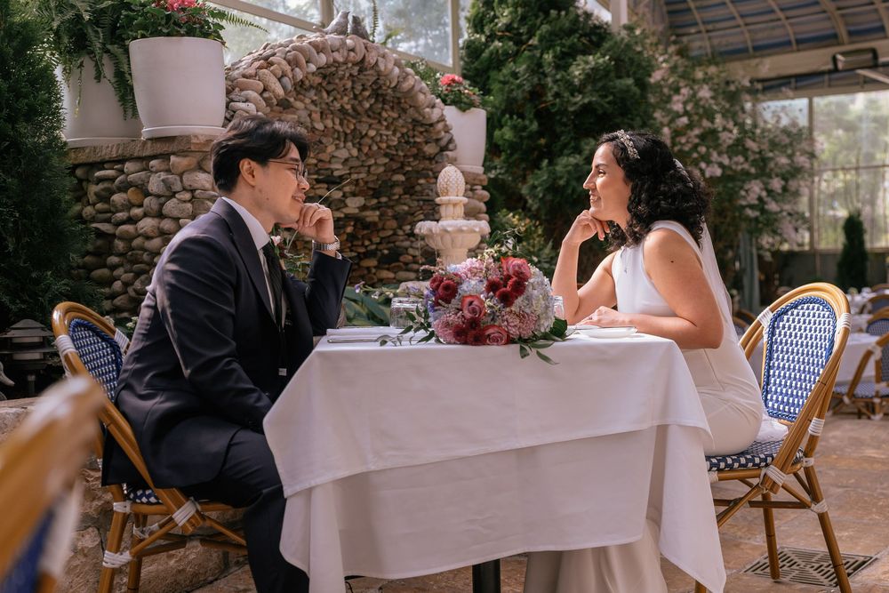 Couple in wedding attire sitting at a white-draped table with floral centerpiece in an elegant garden venue.