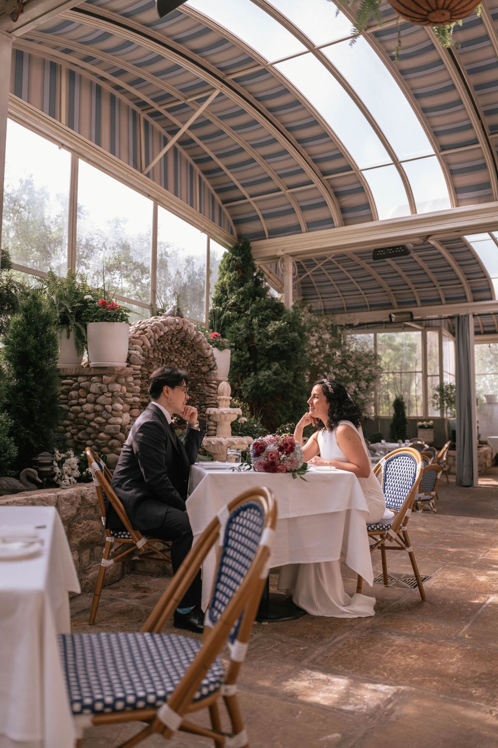 Bride and groom share an intimate moment at a elegantly set table in a sunlit glass conservatory restaurant.