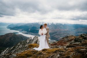Couple embracing in wedding attire on a mountain summit with stunning lake and snow-capped peaks backdrop in New Zealand.
