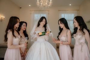 Bride in white gown pours champagne with four bridesmaids in blush pink dresses during wedding preparations.