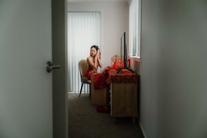 Woman in red dress sitting at vanity mirror doing makeup in softly lit bedroom.