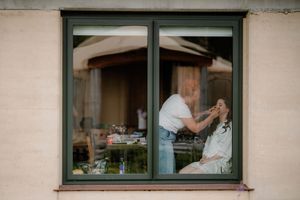 A makeup artist applies makeup to a bride through a window, captured in a candid wedding preparation photo.